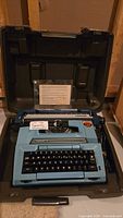 Turquoise Smith Corona Coronet XL Coronamatic typewriter with black keyboard inside open hard case, electric cord visible.