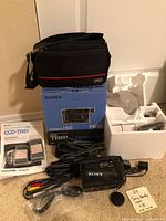 Items including Sony Handycam CCD-TR81 with original box, camera bag, cords, and tapes organized on floor.