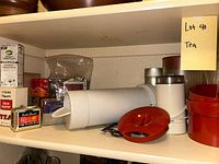 Shelf with white and red plastic pitchers, stacking red bowls, and boxed tea mixes. Items stored in pantry, labeled 'Lot 40 Tea'.