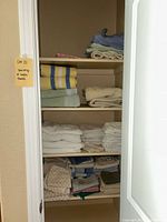 Shelf with folded bath towels and other linens including beige, blue, and white towels with bath mats and shower liner visible