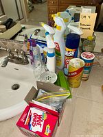 Wide view of the mostly spray bottles and cleaning items on bathroom countertop near sink with labels visible
