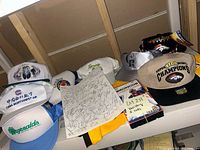 Photo showing multiple baseball caps arranged in two rows with various sports logos including Super Bowl XXXIII Denver Broncos Champions, Colorado Rockies, and others. Signed paper is visible laid flat on the table.
