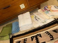 Various folded linens including green lace edged fabrics, blue napkins, white towels, floral patterned cloths and an Indigenous style woven rug on carpeted floor beside wooden dresser.