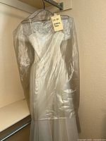 Vintage white wedding dress covered with a clear plastic garment bag hanging on a hanger, showing the fitted silhouette and lace bodice detail through the bag.
