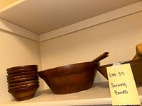 Large wooden serving bowl with two wooden spoons and a stack of 8 small wooden bowls on a shelf, showing the set contents and natural wood tones.