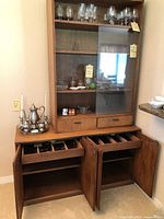 Wide view of wooden china cabinet showing upper glass-front shelves with glassware and two drawers, and lower cabinet with open doors revealing shelves and silverware storage.
