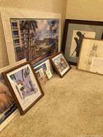 Artworks arranged on carpeted floor and against wall showing variety of framed and unframed pieces