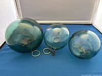 Front view of three glass fishing floats displaying size differences, color, and attached metal rings/chains