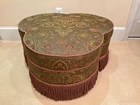 Front view of kidney-shaped ottoman displaying brocade fabric, trim and fringe