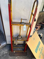 Front view of yellow metal hand truck showing frame, red side supports, strap, toe plate, and wheels
