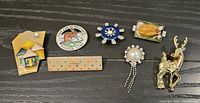 Seven brooches on dark surface including reindeer, pearl tassel, turkey, gear-flower, Anchorage souvenir, enamel house, beige dot rectangle