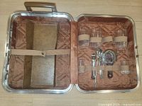 Open briefcase showing six clear ribbed-bottom glass tumblers and five metal bar tools inside quilted brown lining.