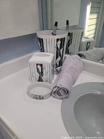 Modern black and white striped ceramic bathroom accessories including toothbrush holder, soap dish, tumbler, and gray towel on a bathroom sink counter.