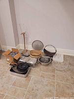 Photo of chafing dishes, pans, woven baskets, and metal bowl arranged on floor against wall.