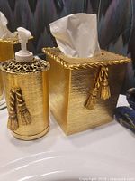 Gold soap dispenser and gold Kleenex box cover with tassel decorations on a bathroom sink.
