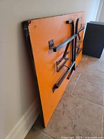 Orange folding table top leaning against wall showing underside and legs folded