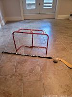 small red hockey goal net on tiled floor with Sher-Wood hockey stick and ball