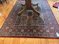 Photo showing full view of Ikea area rug under a wooden table on hardwood flooring. The rug features a traditional red border with floral and geometric detailing.
