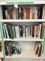 Three white shelves filled with a mixture of books covering novels, gardening, home decor, and golf topics.