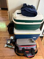 Full view of all items in the camping lot stacked together on wooden floor, showing two hard coolers (green/white and blue/white), thermal soft cooler bag, two small backpacks, and a folded navy blue blanket on top.