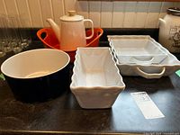 Full view of all items on black countertop including white casserole dishes, orange serving dish, Ikea bowl, and teapot.