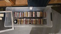 Clear plastic storage bin with approximately 70 to 80 assorted music CDs inside, shown from above.