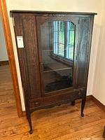 Front and angled views of the wooden china cabinet with glass door showing the general condition, design, and size in a room with wood floor and window reflection