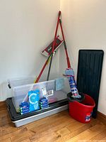 Photo showing the Rubbermaid clear plastic storage totes, mop heads, mop bucket, mop handles, and assorted cleaning supplies grouped together in a corner.