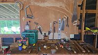 Overview of hand tools arranged on a wooden surface with pegboard background, including saws, hammers, screwdrivers, spirit level, pliers, paint scraper, and the Craftsman drill.