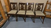 Four wooden dining chairs with harp design on backrests and fabric padded seats arranged side by side.