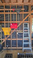 Full view of garage tools including yellow plastic snow shovel, red metal snow shovel, metal leaf rake, wooden broom, garden tool, and aluminum ladder against a wood frame wall.