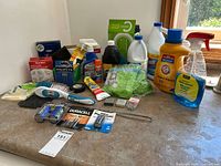 Overview photo of household cleaners, batteries, smoke detector in box, cleaning tools all on counter surface with window background.