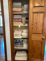 Closet with multiple shelves of folded linens including towels, sheets, table linens, and lace curtains in stacks