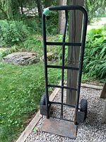 Photo of the entire black metal hand truck dolly standing upright outside on gravel surface, showing height and structure.