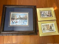 Three framed winter-themed prints arranged on a wooden floor: one large print with ice skating scene and two smaller prints showing children playing in snow.