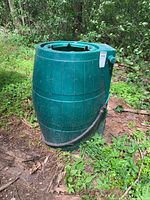Green resin rain barrel standing outdoors with no lid, showing cut top edge and dirt on surface.