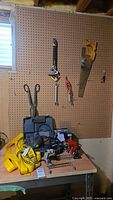 Photo of assorted tools on workbench and pegboard including Mastercraft sander in blue case, hand saw with yellow handle, pliers, clamps, jumper cables, and tow straps.