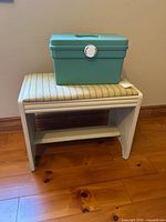 Green plastic sewing box closed and placed on top of a white wooden stool with striped cushioned seat