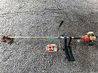 Full view of Stihl FS 90V gas powered weed trimmer laid on gravel with harness strap and spool of replacement trimmer line beside it.