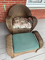 Front angled view of the brown wicker armchair with floral cushion and the matching stool with a green cushion, placed on outdoor decking in front of a brick wall.