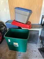 Stacked plastic storage bins in various colors and sizes on a concrete floor against a wood panel wall. The bins include several different colors and shapes showing general wear.