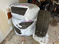 Four snow tires wrapped in plastic, stacked and one leaning against wall showing winter tread