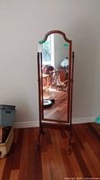 Full view of cheval mirror standing on hardwood floor near wall, showing mirror surface and wooden frame with finials.