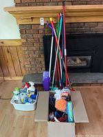 Lot of cleaning supplies including basket with cleaning fluids, cardboard box with gloves and brushes, standing mops and brooms against fireplace.