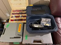 Overview of Dremel 3000 rotary tool with power cord, plastic accessory box, wooden router bit case, and grey plastic case with Brad Nailer inside.
