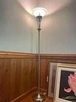 Full view of the floor lamp standing next to wood paneling and framed artwork showing the lamp's metal pole and base and overall height.