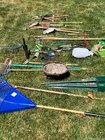 Garden tools spread out on grass showing a mix of handheld and long-handle tools, some metal, plastic, and wood, used and broken.