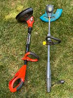 Two string trimmers lying on grass, a red and black Black & Decker model on the left with battery slot and a black Grasshog model on the right with blue trimming head.