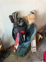 Black TaylorMade golf bag loaded with several golf clubs and a green protective cover nearby. Cardboard boxes and other household items in background.