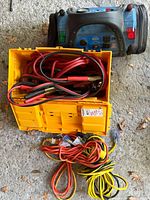 Photo showing yellow storage case with heavy duty red jumper cables and clamps coiled inside, additional orange and yellow extension cords coiled on ground.